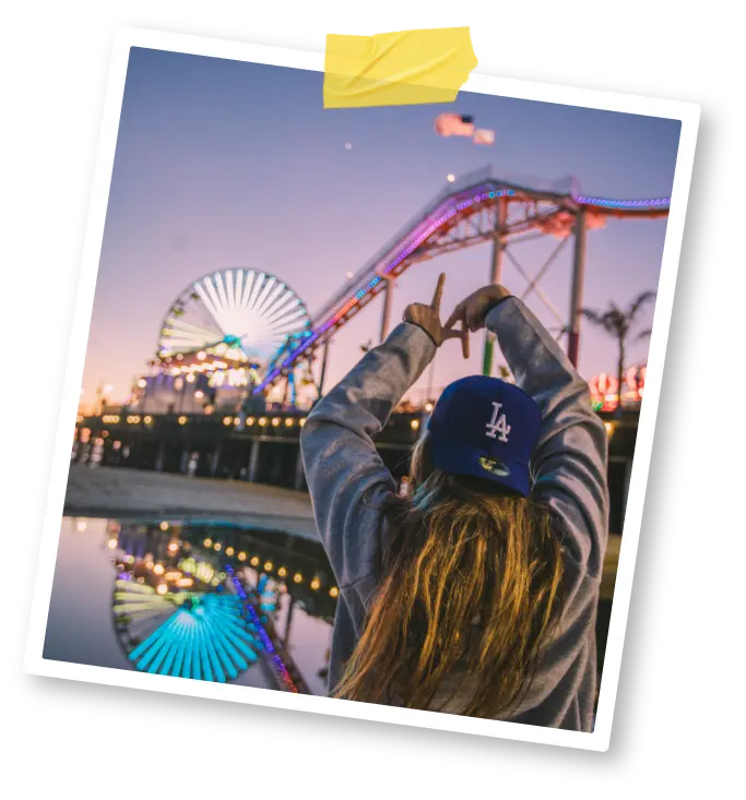 Young woman in a baseball hat makes the letters LA with her fingers against a backdrop of a boardwalk at twilight