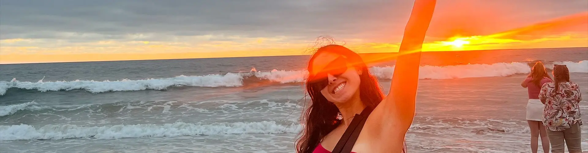 Girl with sunglasses standing with her arm raised on a beach with waves and sunset in the background